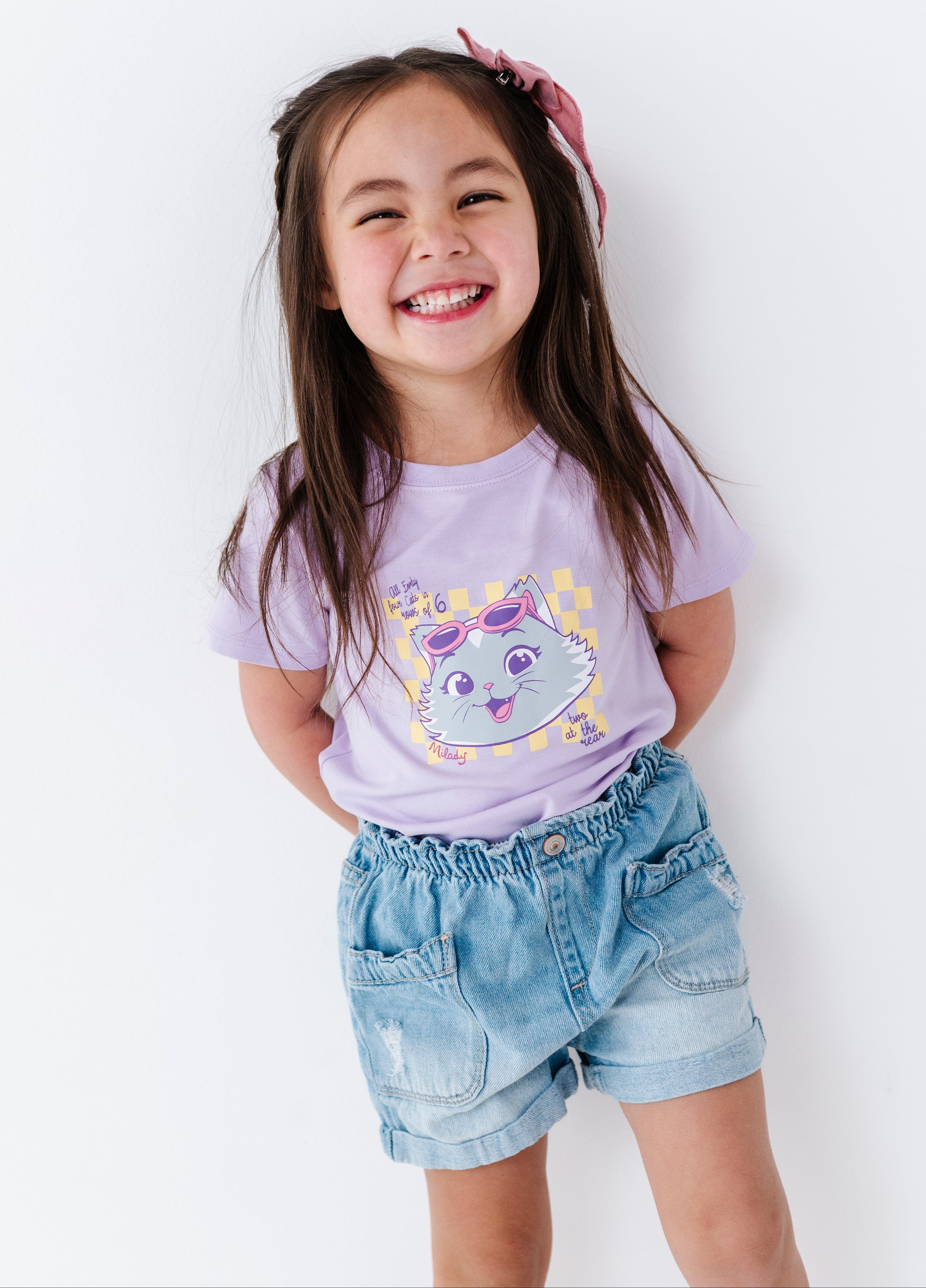 Young girl wearing a purple 44 Cats t-shirt with a cartoon design and denim shorts, standing against a white background.
